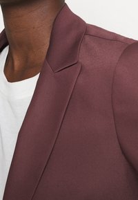 Burgundowy żakiet wykonany z gładkiego materiału, z wykładanym kołnierzykiem, strukturalnymi ramionami oraz schludnym, dopasowanym krojem. Biała koszula pod spodem.