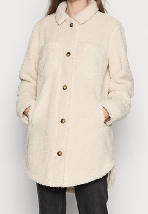 Persona que lleva una chaqueta sherpa de color crema con botones marrones, cuello, mangas largas y bolsillos, combinada con pantalones negros.