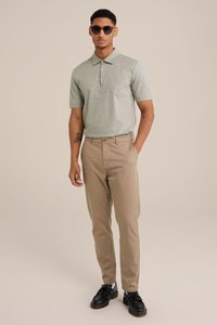 Šedá polokošile s krátkým rukávem a kapsou, světle hnědé kalhoty a černé boty. Model má na sobě sluneční brýle; neutrální pozadí.