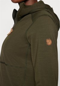 Tume roheline kapuutsiga särk, millel on tõmblukuga esiosa, tekstureeritud kangas ja väike pruun emblem vasakul varrukal. Õmblused on nähtavad.