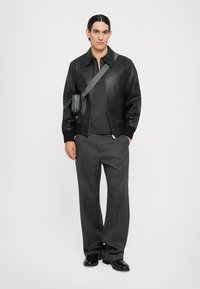 Schwarze Lederbomberjacke mit gerippten Bündchen, grau gestrickter Pullover und graue weite Hose. Eine kleine schwarze Umhängetasche wird getragen.