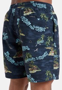Navy swim shorts featuring tropical prints with palm trees and text accents. They have an elastic waistband and a rear pocket with a snap closure.