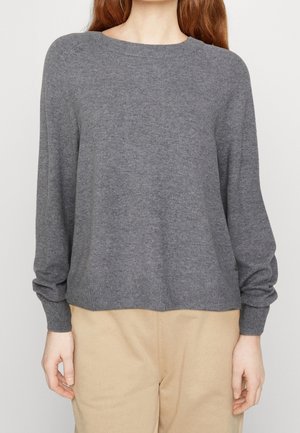 Femme portant un pull gris uni à manches longues et un pantalon beige, debout avec les mains détendues le long du corps sur un fond uni.