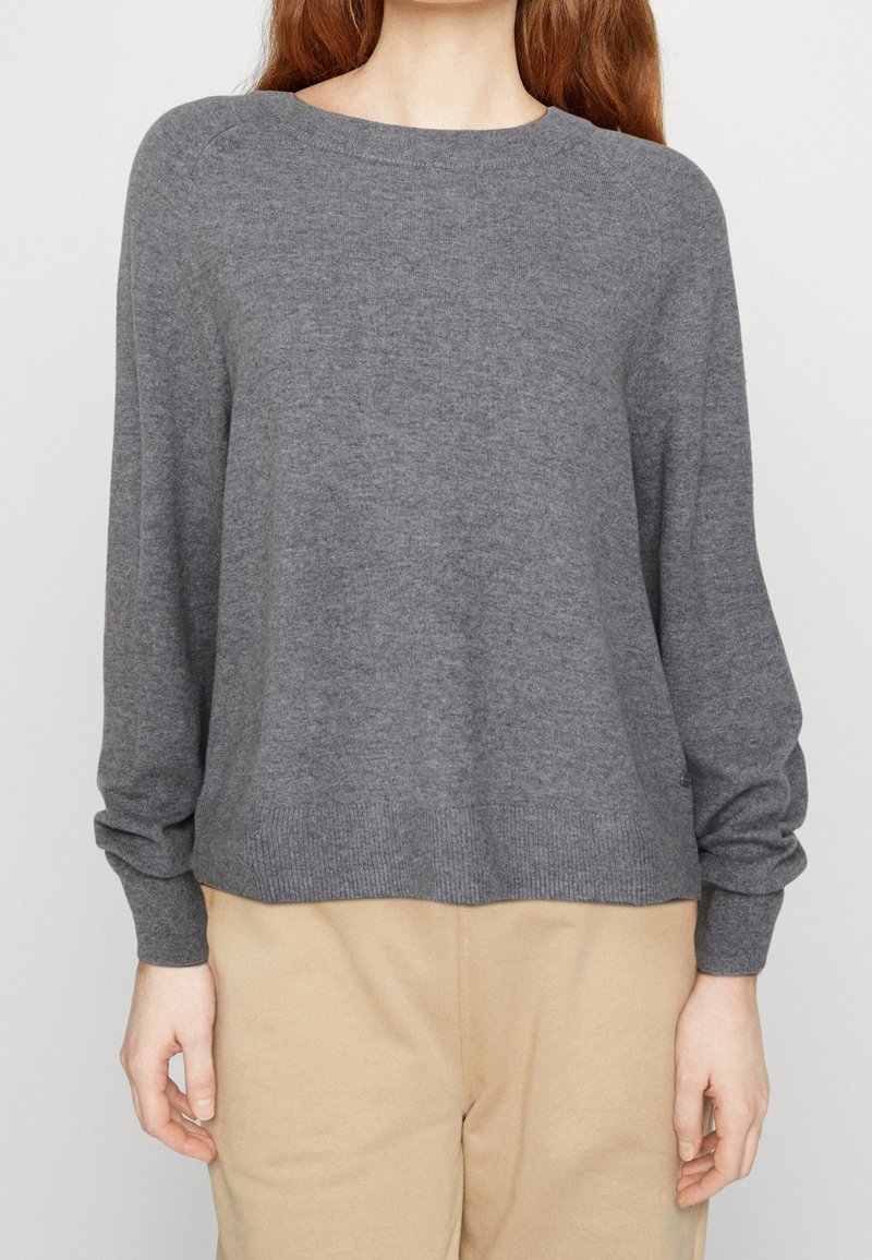 Femme portant un pull gris uni à manches longues et un pantalon beige, debout avec les mains détendues le long du corps sur un fond uni.