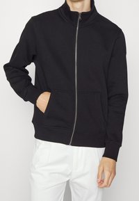Svart zip-up sweatshirt med hög krage, två framfickor och ribbade ärmslut. Mjuk material med en jämn yta, visad med vita byxor.