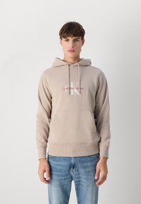 Felpa beige realizzata in tessuto liscio, dotata di tasca kangaroo frontale, cappuccio con cordino e logo con testo rosso sul petto.