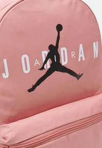 Zaino in tessuto rosa con silhouette del logo Jordan nera e testo "JORDAN" in bianco. Include una tasca anteriore con zip e materiale testurizzato.
