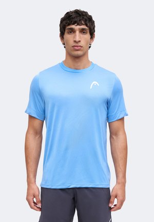 Jonge man met kort krullend haar, die een hemelsblauw sportshirt met korte mouwen en donkere shorts draagt, staand tegen een effen lichte achtergrond.