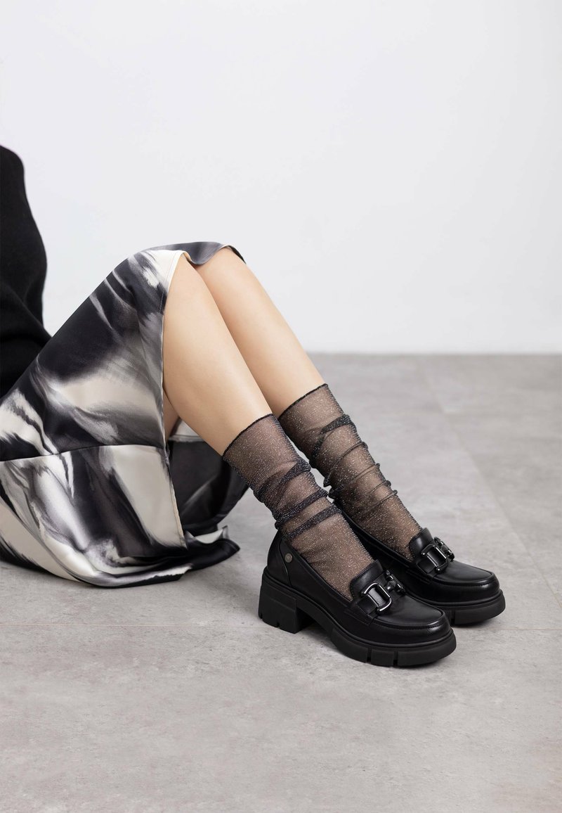 Zapatos de plataforma gruesos y negros con un detalle de hebilla, combinados con calcetas negras transparentes y texturizadas. El fondo presenta una falda de satén en tonos grises.