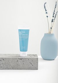Benton PHA Peeling Gel i en mjuk blå tub med vit text, placerad på en strukturerad grå stensockel med en ljus vas i bakgrunden.