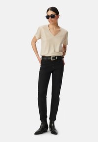Camiseta beige de cuello en V, pantalones ajustados negros, cinturón negro con tachuelas y botines negros. La modelo lleva gafas de sol oscuras y pulsera dorada.