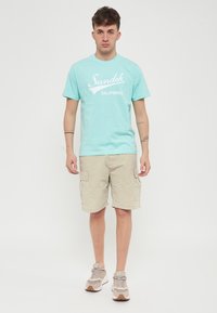 Maglietta azzurra a maniche corte con stampa bianca "Sundek California", abbinata a pantaloni cargo beige e sneakers in tonalità neutre.