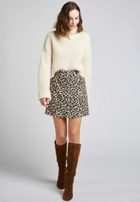 Pull en crème épais, col montant, texture côtelée ; jupe brune à motifs léopard avec des boutons ; bottes en suède brun hautes au design ajusté.