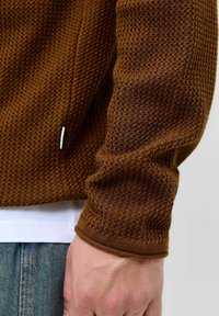 Braun melierter Strickpullover mit geripptem Bündchen, enganliegendem Design und dezenter Musterung. Weißes Etikett an der Seitennaht sichtbar.