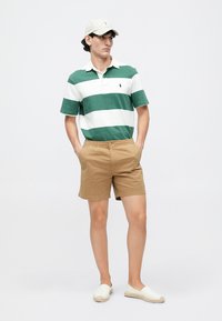 Zaļi un baltā svītraina polo krekls, khaki šorti un gaiši ievelkamās kurpes. Galvā ir cepure. Apģērbs ir ērti un neformāli stilizēts.