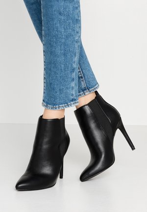 Beine in blauen Jeans, die schwarze spitze Lederknöchelstiefel mit hohen Stiletto-Absätzen auf einem weißen Boden tragen.