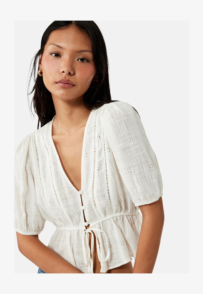 Joven mujer con cabello largo y oscuro que lleva una blusa blanca texturizada con mangas abullonadas y botones delanteros atados en la cintura.