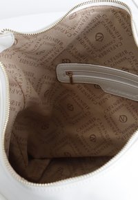 Interior de un bolso blanco con un forro liso que presenta el logotipo repetido "CAFE NOIR" en marrón claro. Incluye un bolsillo con cremallera y una cremallera dorada.