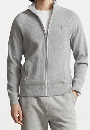 Muž v lehce šedé texturované bundě na zip s žebrovanými manžetami a pasem, přes bílou košili, doplněný o odpovídající šedé tepláky.
