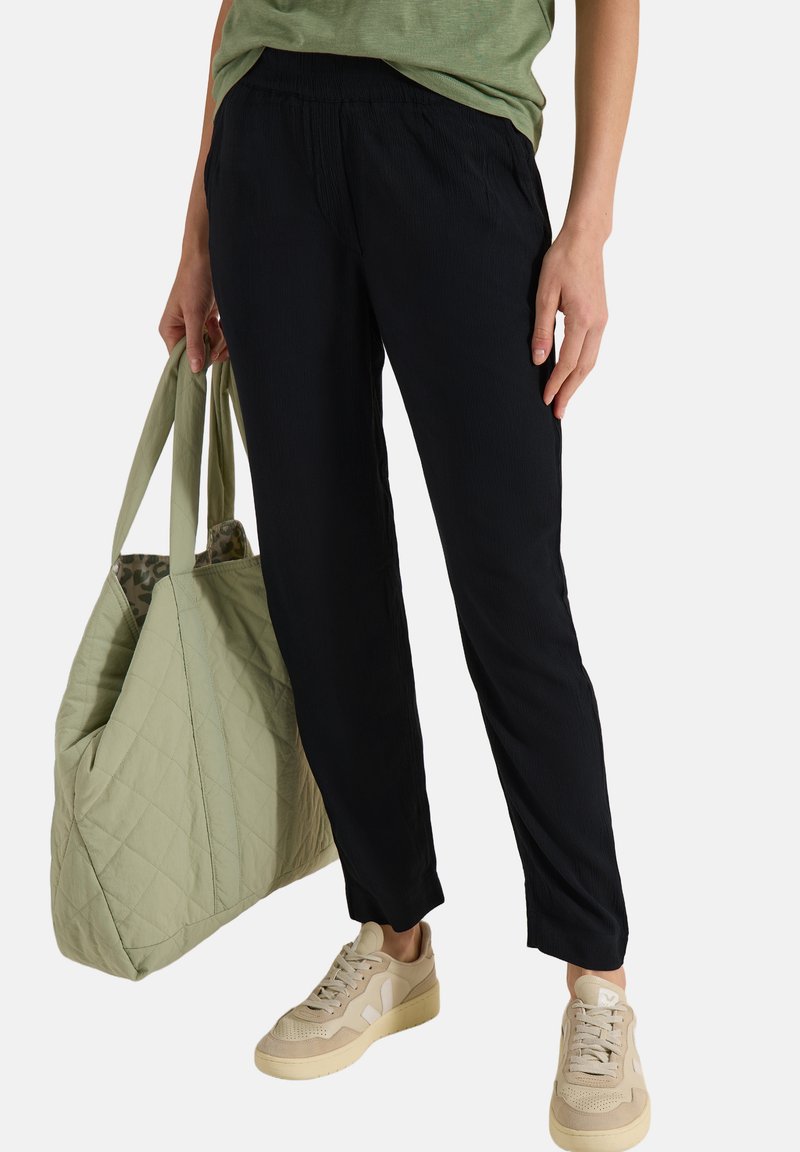 Persona que lleva pantalones negros ajustados, zapatillas beige, top verde, sosteniendo un gran bolso tote verde acolchado al lado.