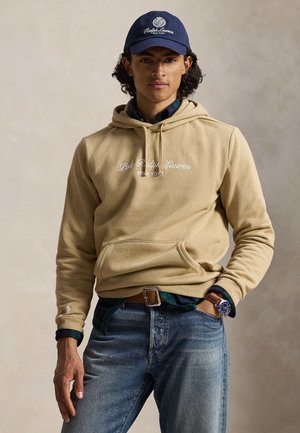 Junger Mann trägt einen beigen Polo Ralph Lauren Hoodie, blaue Jeans, dunkelblaue Mütze und Uhr, steht mit einer Hand in der Tasche vor einfarbigem Hintergrund.