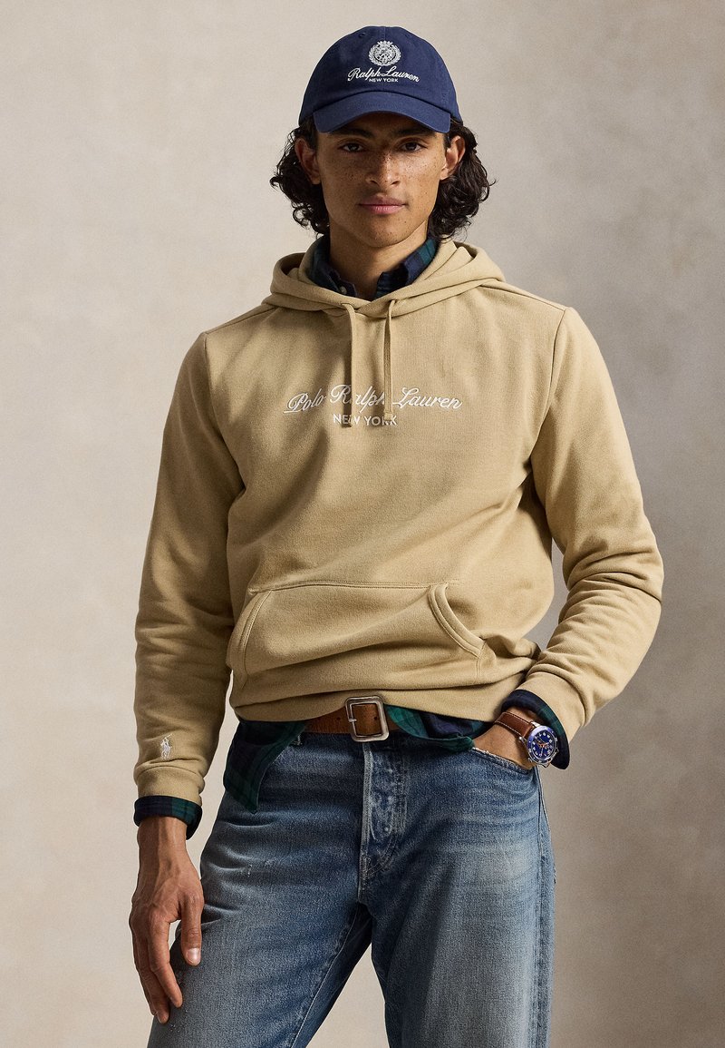 Νέος άνδρας φοράει μπεζ φούτερ Polo Ralph Lauren, μπλε τζιν, σκούφο ναυτικού χρώματος και ρολόι, στέκεται με το ένα χέρι στην τσέπη μπροστά σε απλό φόντο.