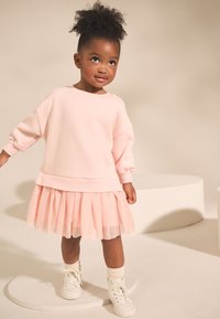 Világos rózsaszín pulóver ruha hosszú ujjakkal, réteges tüll szoknyával és bordázott mandzsettákkal, fehér magas szárú tornacipővel párosítva.