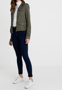 Kvinna som bär en olivgrön jacka, en vit ribbad polo, mörkblå skinny jeans och vita sneakers mot en enkel bakgrund.