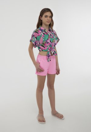 Camisetta corta a maniche corte con un motivo di foglie colorato, abbinata a pantaloni corti rosa chiaro e infradito neutri. Texture lisce in tutto il capo.