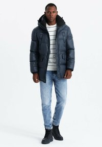 Giacca a vento blu navy con chiusura a zip e bottoni automatici, dotata di ampie tasche frontali e cappuccio. Indossata sopra un maglione a righe e jeans chiari.