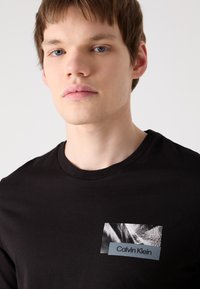Melna kokvilnas t-krekls ar kvadrāta grafikas plāksteri, uz kura ir strukturēta monochromā bilde un zila etiķete ar uzrakstu "Calvin Klein."