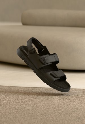 Zwarte sandalen met twee verstelbare bandjes, een rubberen zool met structuur, open teenontwerp en geperforeerd voetbed, weergegeven tegen een neutrale achtergrond.