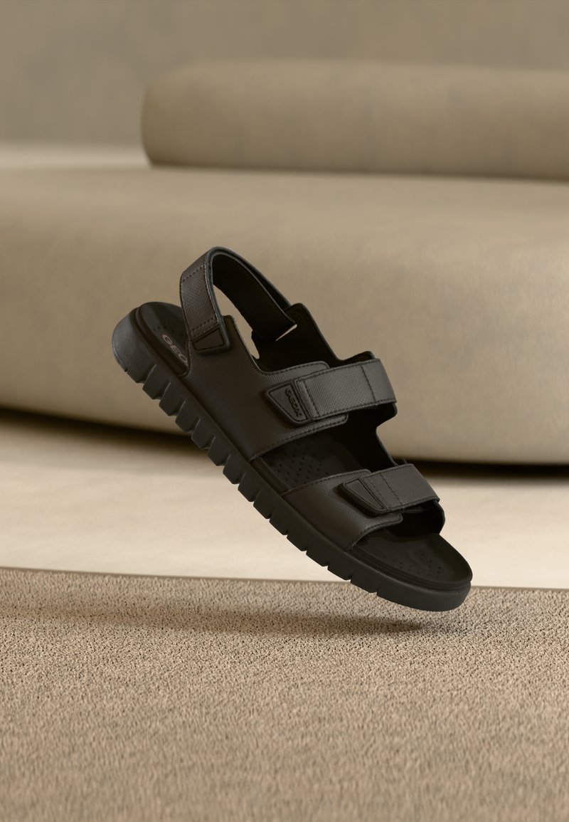Μαύρα σανδάλια με δύο ρυθμιζόμενους ιμάντες, σχέδιο με ελαστική σόλα με υφή, ανοιχτή μύτη και διάτρητη επιφάνεια υποδημάτων, παρουσιασμένα σε ουδέτερο φόντο.