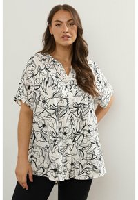Blusa bianca con motivo floreale nero, maniche corte, scollatura a V, bottoni davanti e vestibilità rilassata. Adatta per un look casual.