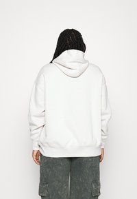 Bílá mikina s kapucí a uvolněným střihem, se stahovací šňůrkou a žebrovanými manžetami, v kombinaci se šedými cargo kalhotami.