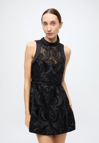 Black sleeveless dress with floral embossed design, sheer mesh upper, and a high collar. Features a defined waist and smooth texture.
