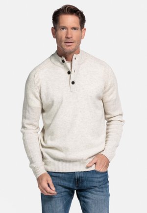 Man in een lichtbeige gebreide trui met knopen en een spijkerbroek, staande tegen een plain witte achtergrond.
