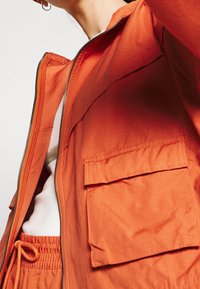 Orange lättviktsjacka med dragkedja framtill, två framfickor och avslappnad passform, kombinerad med en vit topp och matchande shorts med dragsko.