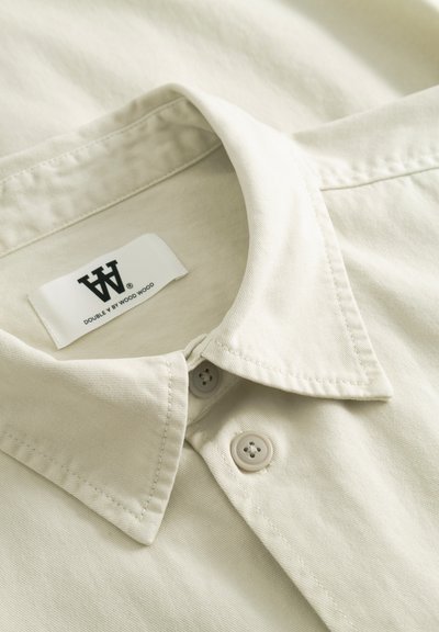 Chemise beige à col avec fermeture par boutons et étiquette de marque visible "Double V by Wood Wood" à l'intérieur du col.