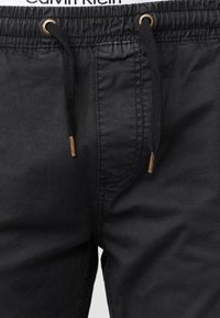 Black cotton shorts with a drawstring waistband, featuring gold-tone eyelets and a smooth texture. Minimalist design with no visible patterns.