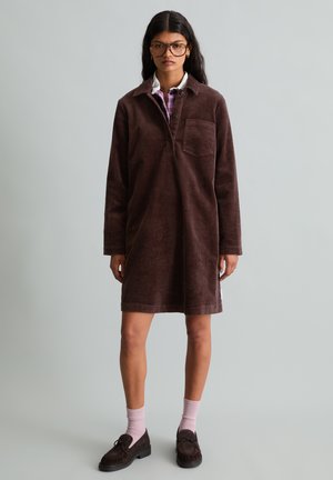 Jonge vrouw met lang donker haar die grote bril draagt, een bruine ribfluwelen jurk, roze sokken en donkere loafers tegen een effen lichtgrijze achtergrond.