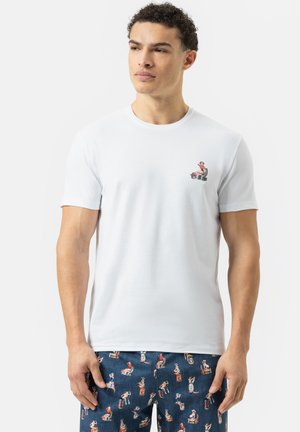 Weißes Baumwoll-T-Shirt mit einem kleinen grafischen Design auf der linken Brust. Kombiniert mit navyblauen Hosen, die bunte Muster von Figuren im Vintage-Stil zeigen.