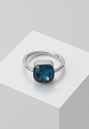 Srebrny pierścionek z dużym kwadratowym, głęboko niebieskim, fasetowanym kamieniem osadzonym na środku, prezentowany na białym geometrycznym bloku.