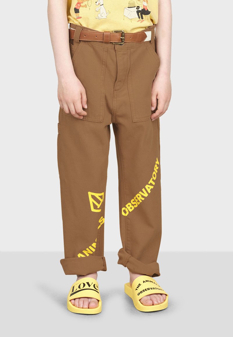 Pantalons cargo marron avec des motifs graphiques jaunes, dotés d'une coupe décontractée, de poches doubles et d'une ceinture marron. Associés à des sandales à enfiler jaunes.