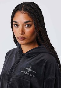 Sudadera con cremallera de terciopelo negro que presenta una capucha, bolsillos frontales y un acento de logo adornado con joyas plateadas en el área del pecho izquierdo.