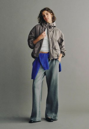 Young woman stands with one hand in pocket, wearing gray jacket, white crop top, gray wide pants, blue sweater tied around waist, and black shoes.