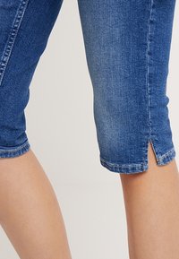 Capri in denim di blu medio, con un design aderente, spacchi sull'orlo e dettagli di cucitura. Il materiale ha una superficie leggermente tessurizzata.