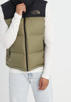 Osoba nosząca oliwkowo-zieloną i czarną bezrękawnik puchowy z logo The North Face, białą bluzkę z długim rękawem oraz jasnoniebieskie dżinsy.