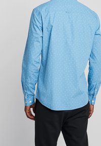 Muž v světle modré košili s dlouhým rukávem a malými bílými vzory, doplněné černými kalhotami, viděný zezadu.