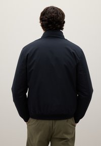 Uomo con capelli ricci e castani indossa una giacca nera e pantaloni verde oliva, in piedi con le mani in tasca, di fronte a un muro beige uniforme.